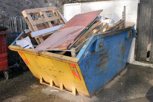 A large, open-top metal skip in a yellow and blue finish positioned on a concrete surface outdoors, filled with a variety of waste materials including wooden pallets, metal sheets, and construction debris. The wooden pallets, visible on top of the pile, are light-colored with horizontal slats, some partially broken or leaning. The metal sheets are rusty with a reddish-brown tint, and the debris appears haphazardly loaded, indicating disposal or clearing activity. In the background, a white wall and a black wooden fence delineate the area, with some tools or storage items partially visible to the left. The scene is illuminated by natural daylight, casting minimal shadows. This setup is typical of commercial waste disposal operations often conducted by companies like Commercial Waste Croydon in the Croydon area, providing rubbish removal services within the local postcode district.