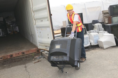 A female worker wearing a yellow safety helmet, high-visibility vest with red and yellow stripes, and gloves is positioned outdoors near an open shipping container on a paved surface. She is holding a portable computer monitor with her right hand, which is attached to a black and gray waste control device or portable unit. Behind her, the interior of the container reveals a cluttered arrangement of various electronic appliances such as old CRT monitors and computer towers, along with other discarded items. In the background, there are some stacked bricks and a white temporary structure, suggesting a waste disposal or collection site. The setting appears to be part of a commercial waste collection operation in Croydon, with clear focus on rubbish removal services and environmentally aligned waste handling practices.