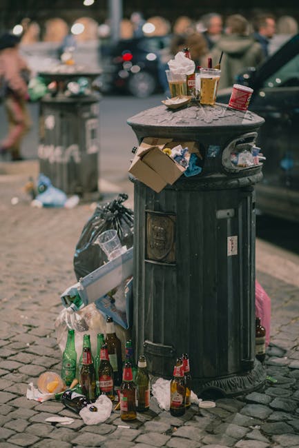 A black cylindrical waste bin, positioned against a large, light-colored, textured stone or concrete wall, is overflowing with various food packaging, wrappers, and takeout containers made of cardboar