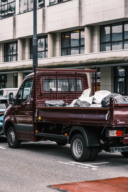 A small dark red flatbed truck parked on an urban street in front of a modern office building with large glass windows and a light-colored facade. The truck's cargo bed is filled with various types of