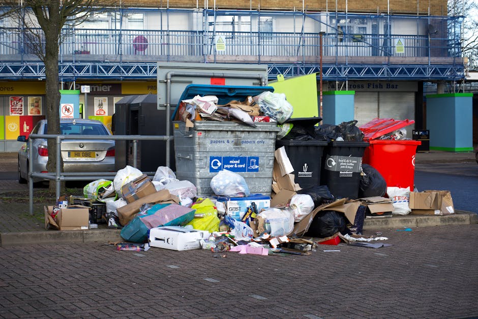 A collection of disorganized cardboard boxes and paper waste piled in front of a white corrugated metal wall, with a prominent blue sign reading 'Recycling Only' featuring a green recycling symbol. Th