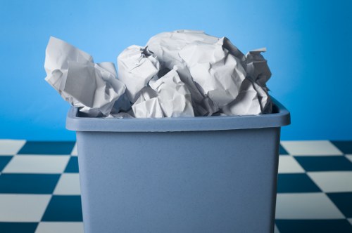 A light blue plastic bin filled with crumpled white paper waste, situated on a checkered black and white tiled surface, with a blue gradient background. The paper appears to be loosely compressed and slightly torn, with some sheets protruding over the top edge of the bin. The bin's surface has a smooth matte finish, and the overall scene is well-lit, emphasizing the texture of the crumpled paper and the clean, simple design of the waste container. This image reflects rubbish disposal or waste collection, aligning with services provided by Commercial Waste Croydon in the Croydon area, which specialises in commercial rubbish removal solutions.